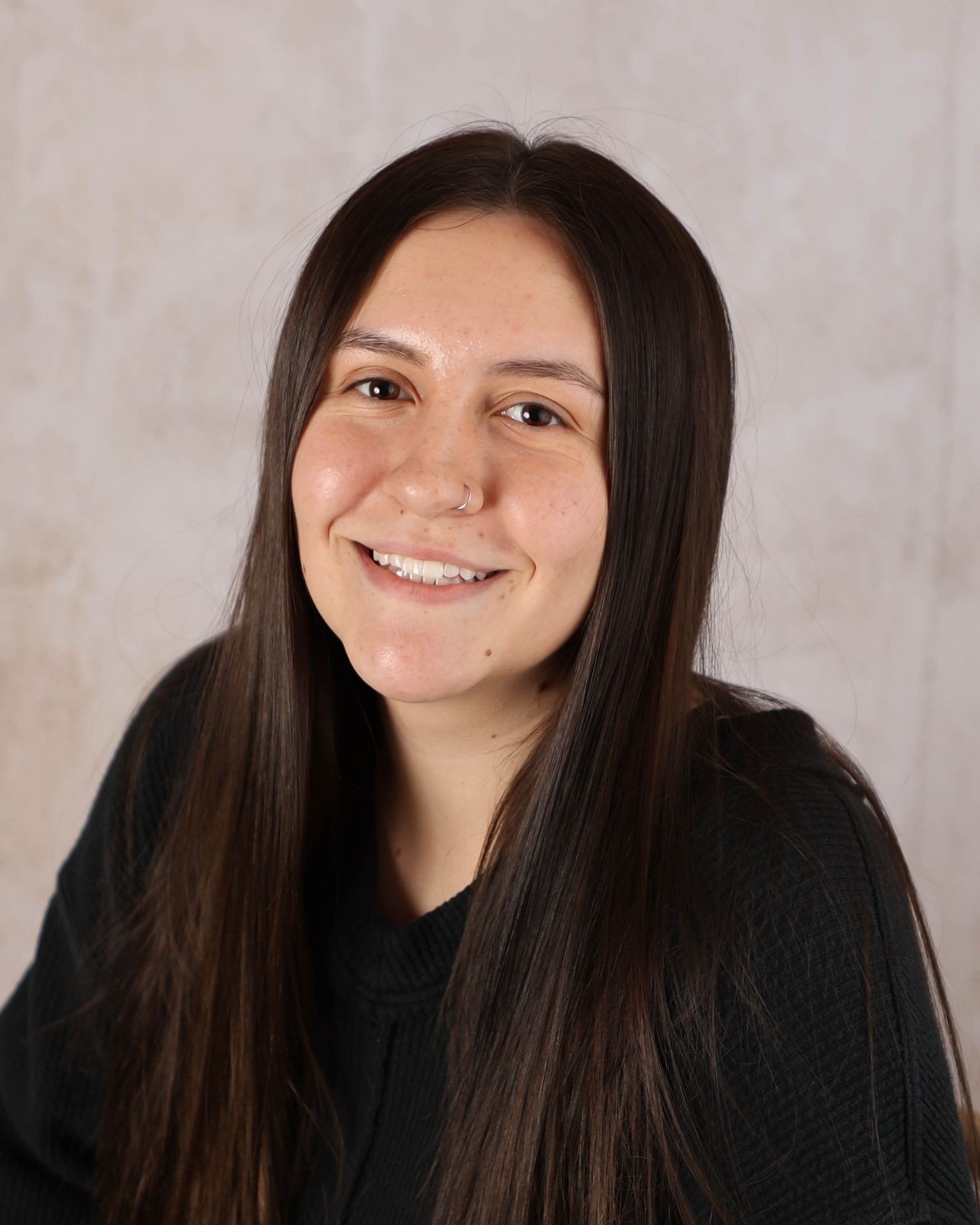 Smiling person with long dark hair and a nose ring in front of a neutral background.
