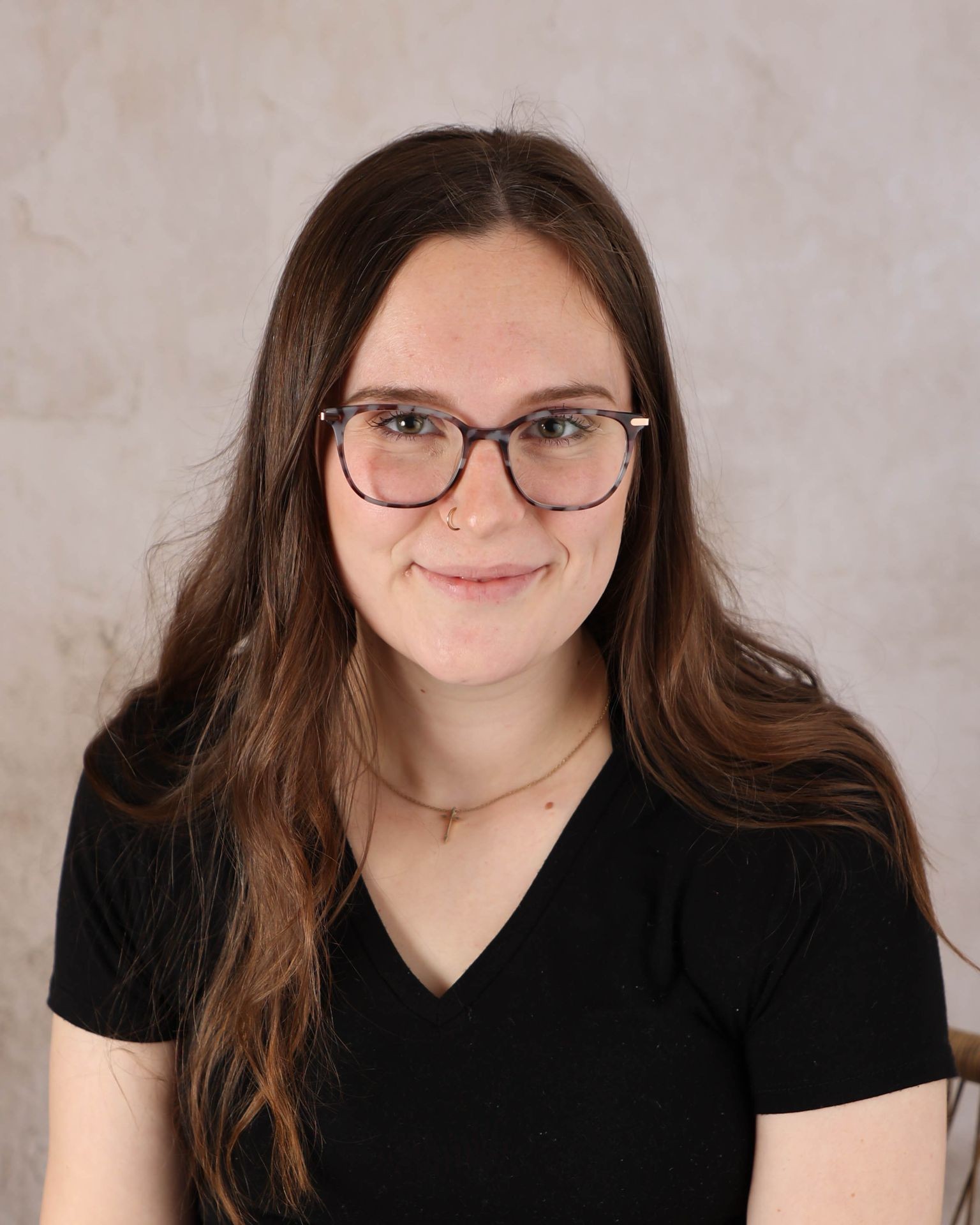 Person with glasses smiling, wearing a black shirt and necklace, against a light background.