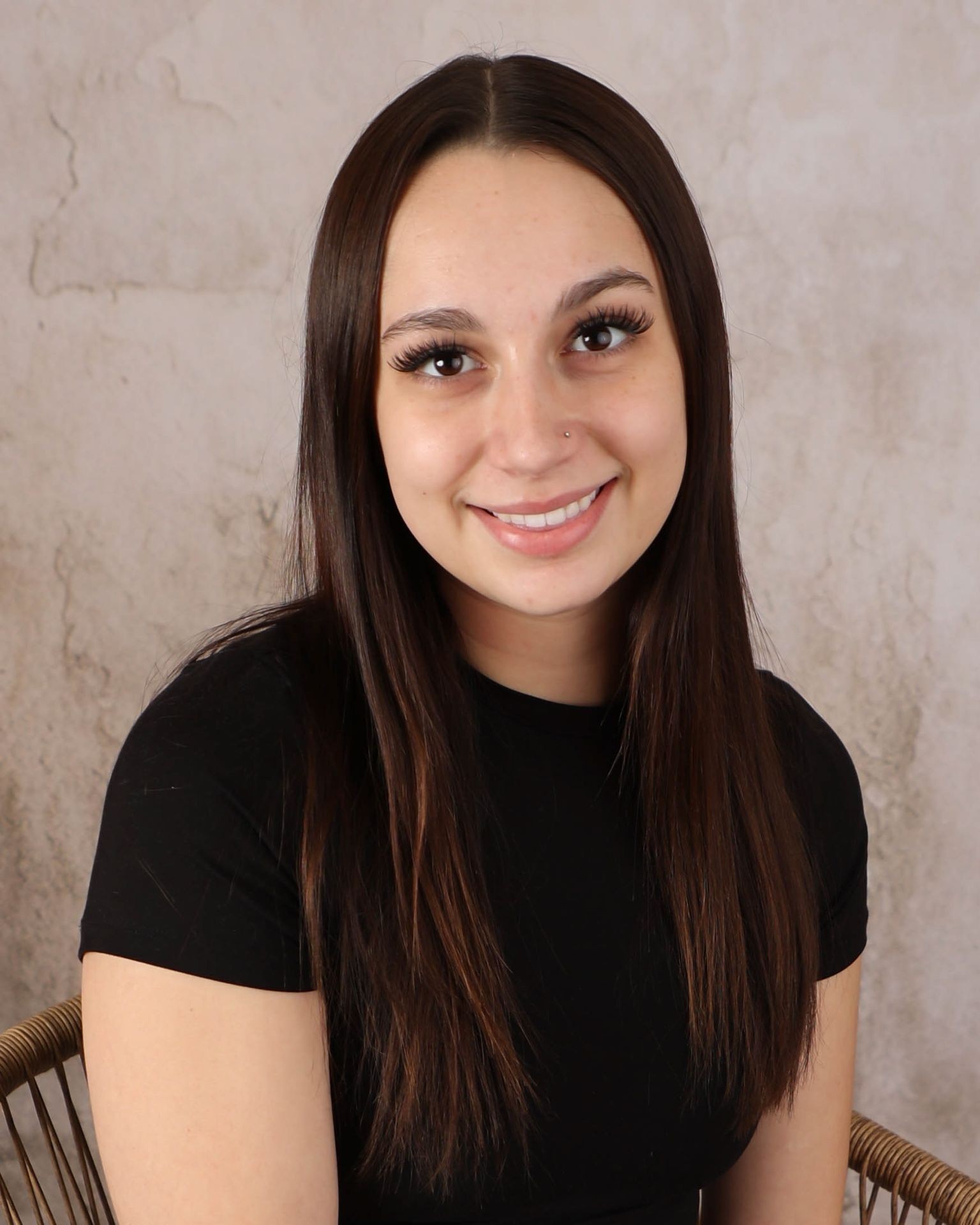 Person with straight, long brown hair wearing a black shirt and seated with a light textured backdrop.