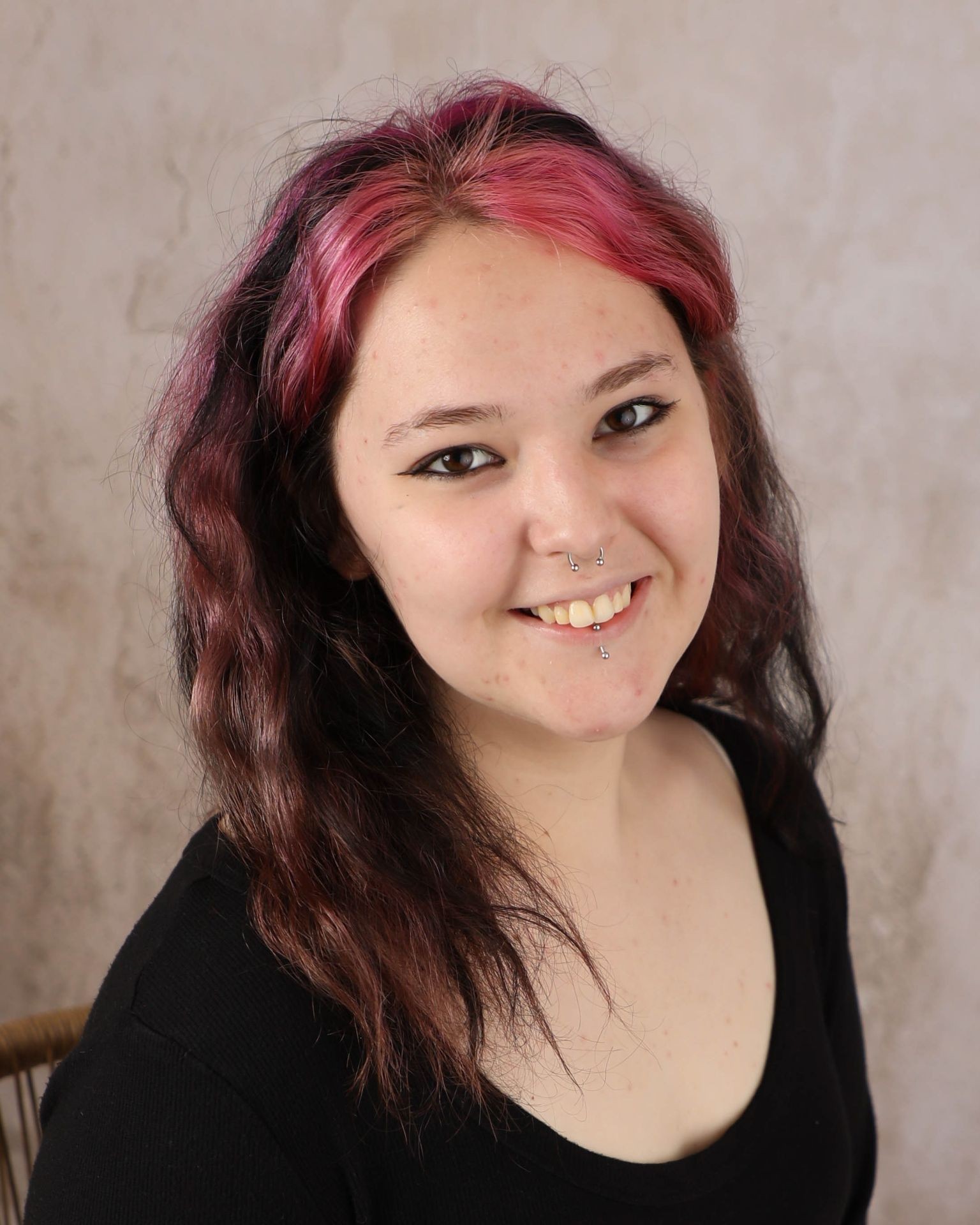 Person with pink hair and piercings smiles against a neutral background.