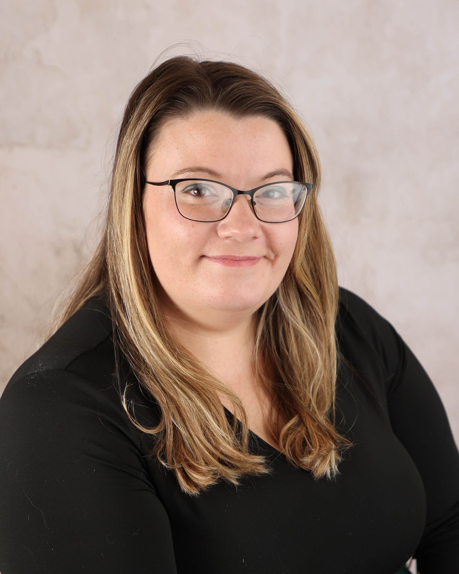 Woman with long hair and glasses smiling, wearing a black top against a light background.
