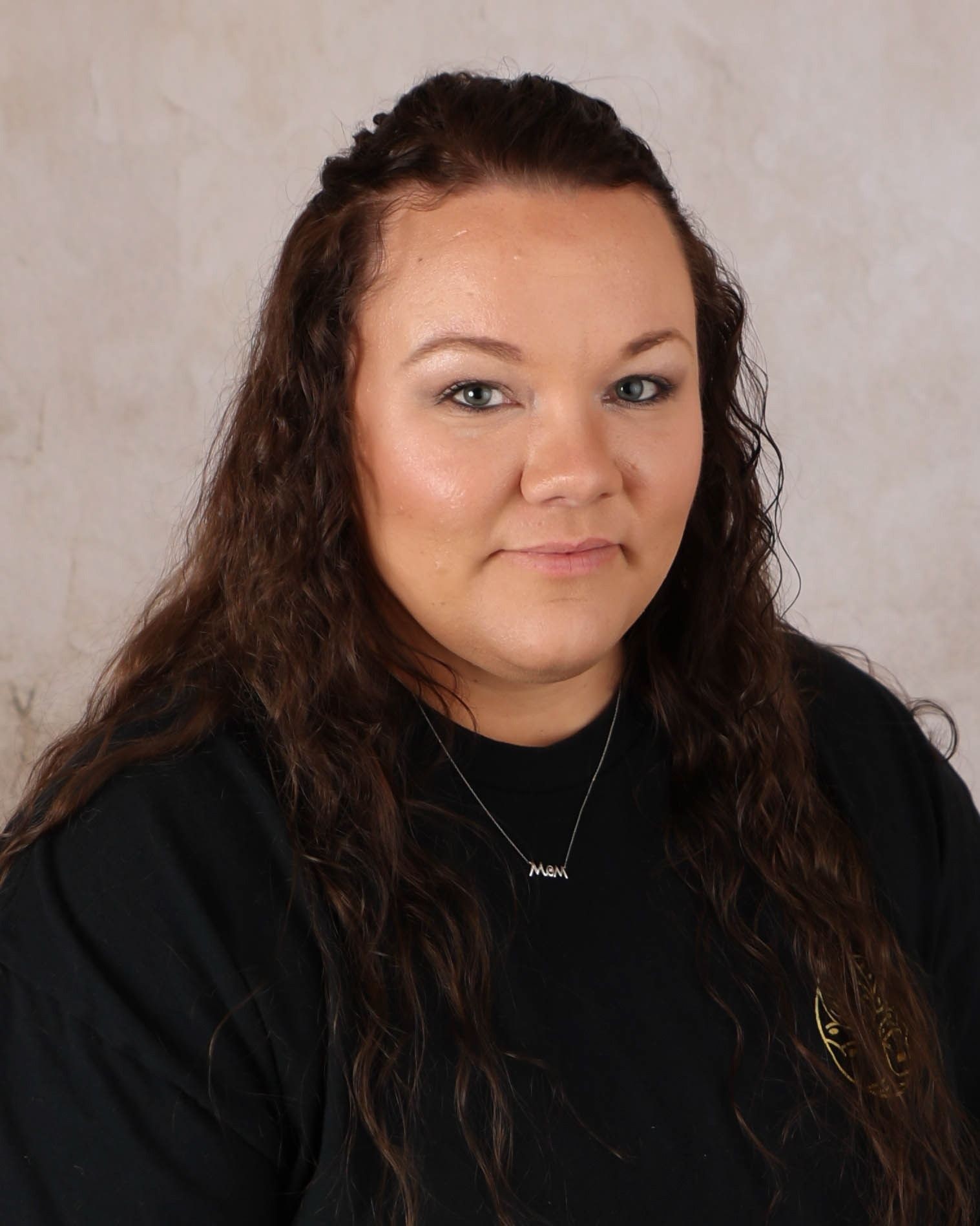 Woman with long curly hair wearing a black shirt and necklace, posing against a neutral background.