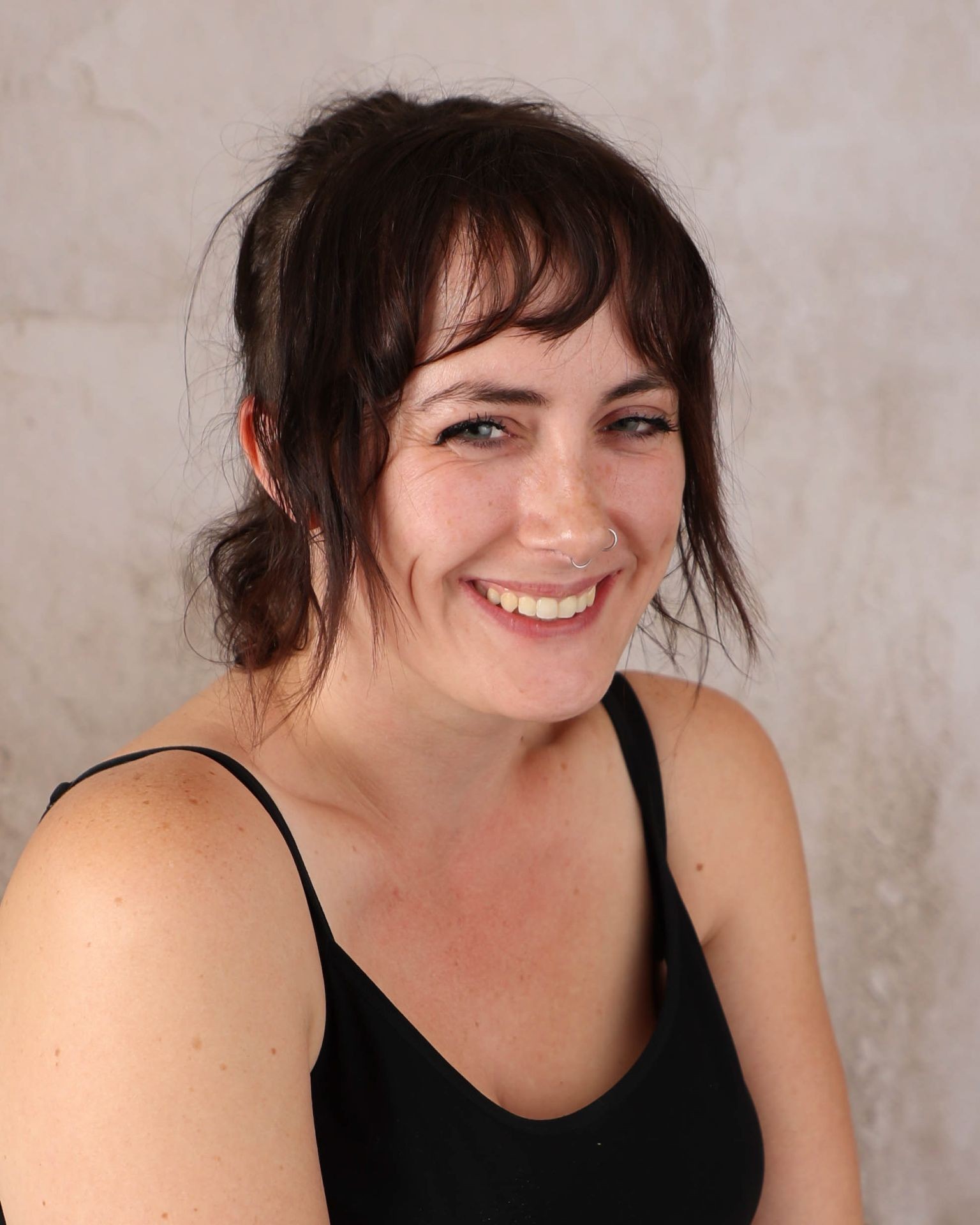 Woman with dark hair and a nose ring smiling in front of a neutral background.