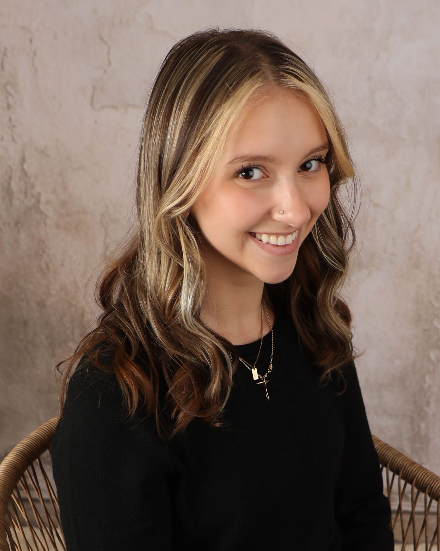 Smiling person with long wavy hair and necklace, seated in wicker chair against textured wall.
