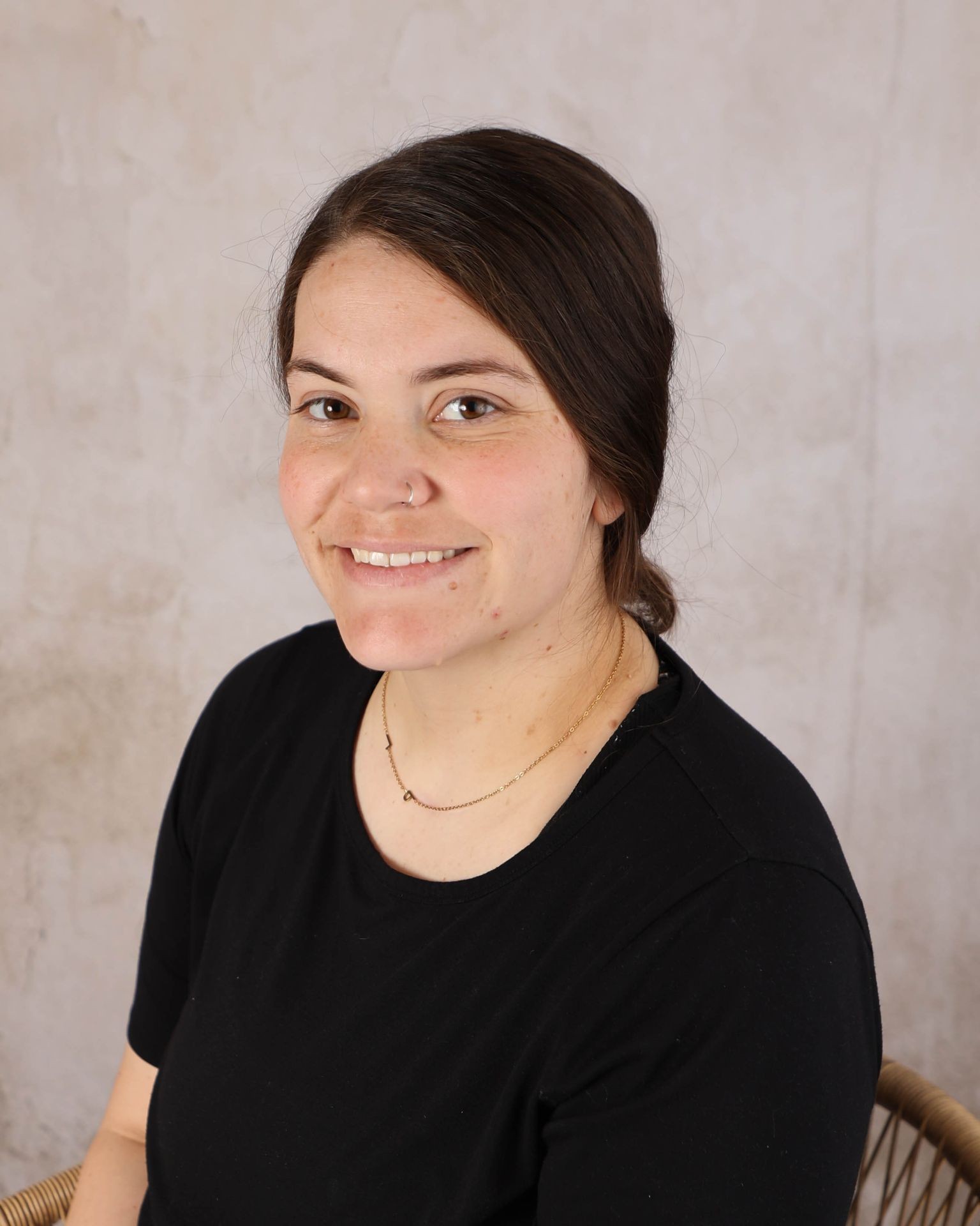 Woman with a nose ring and black shirt smiling against a textured beige background.